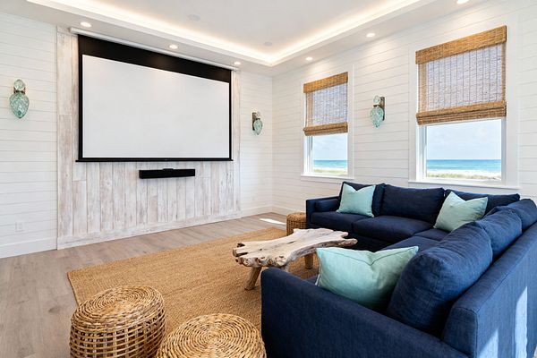 A bright coastal media room features white shiplap walls, a whitewashed plank feature wall with a drop-down projector screen and floating soundbar, and two windows with woven shades overlooking the ocean. A deep navy sectional with pale aqua pillows surrounds a sculptural driftwood coffee table on a large jute rug, with two woven poufs nearby. Recessed and cove lighting softly illuminate the space, complemented by aqua glass wall sconces.