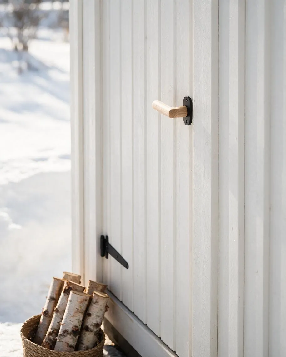 A close-up of a white vertical wood exterior wall or door on a Scandinavian-style cabin, featuring a simple light wood handle and black metal hardware. A woven basket filled with birch logs sits on the snowy ground beside the structure, with soft winter light creating a calm, minimal atmosphere.