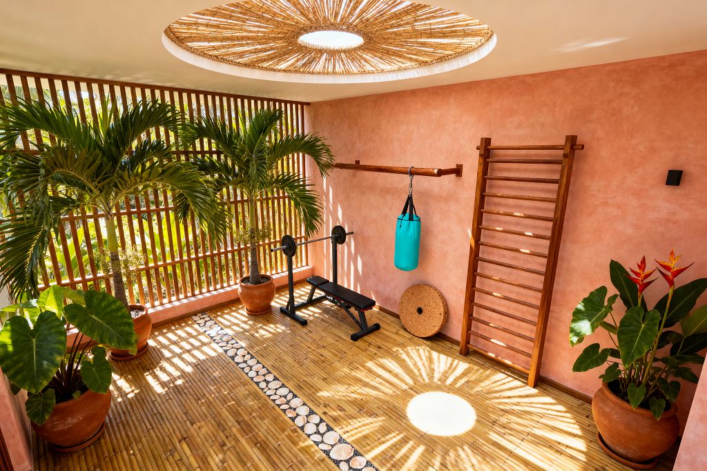 A tropical-inspired home gym with coral stucco walls and a golden bamboo floor features a wood-slat screen opening to greenery. A circular skylight with a woven rattan shade throws a sunburst of light. Equipment includes a black weight bench with barbell, wall-mounted wooden stall bars, a wooden rail with a small turquoise punching bag, and a cork balance disc. Terracotta pots hold palms, elephant-ear leaves, and bird-of-paradise, creating an indoor–outdoor feel.