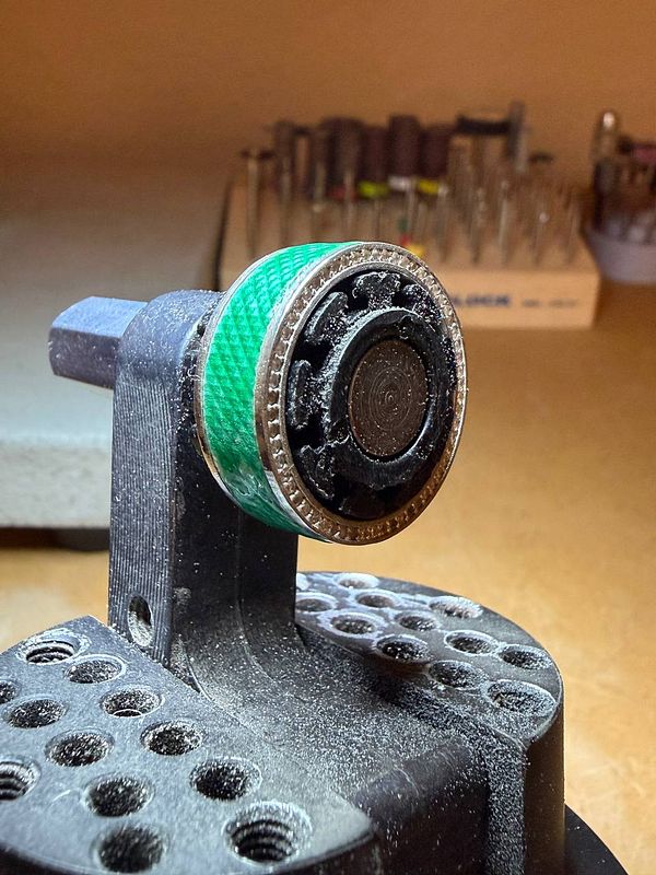 A close-up photo of a person's hand holding a polished metallic ring with intricate arc-shaped inlays against a dark background.
