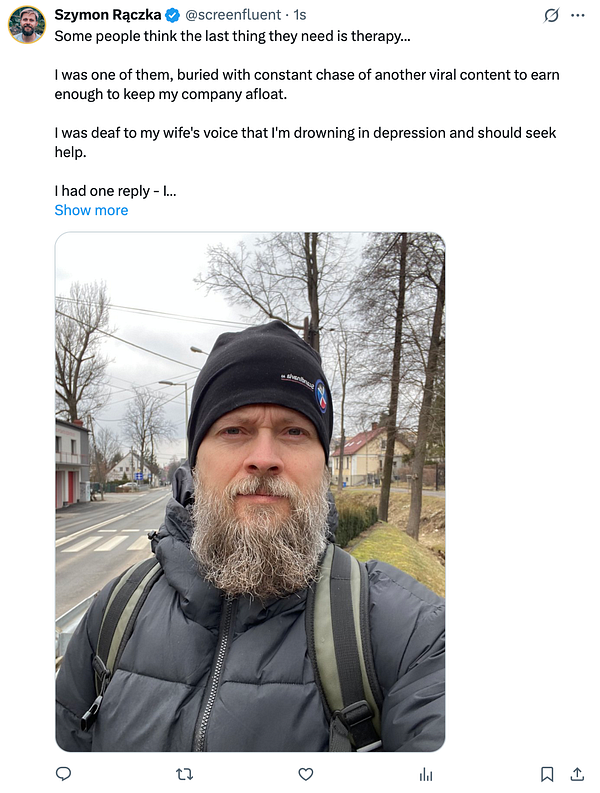 A man with a beard stands outdoors, wearing a black beanie and a puffy jacket, with a street and trees in the background.
