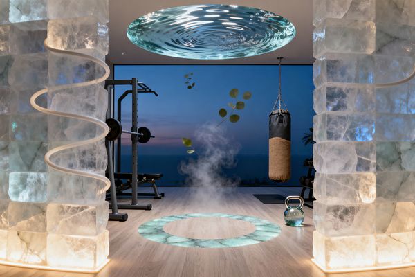 A surreal home gym overlooks the ocean at dusk through a floor‑to‑ceiling window. Two translucent stone block columns glow softly, one wrapped with a sinuous light. A circular, rippled glass feature centers the ceiling above a lit onyx ring in the floor releasing mist. A cork-and-black leather punching bag hangs to the right, with a glass kettlebell nearby and a steel squat rack to the left. Eucalyptus leaves appear to float midair, enhancing the dreamlike scene.
