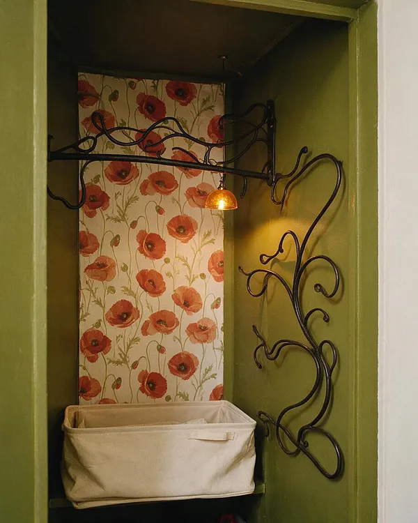A small closet interior painted olive green, featuring black wrought-iron scrollwork along the sides and top, a warm amber pendant light, and a back panel covered in poppy floral wallpaper. A beige fabric storage bin sits on the lower shelf, adding a soft neutral contrast.