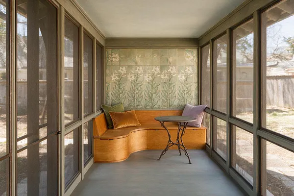 A screened front porch with dark-framed mesh windows on both sides, a curved built-in wooden bench at the back, and a small round metal table in front. The rear wall features decorative floral Art Nouveau tiles in soft green tones, with muted accent pillows adding color.