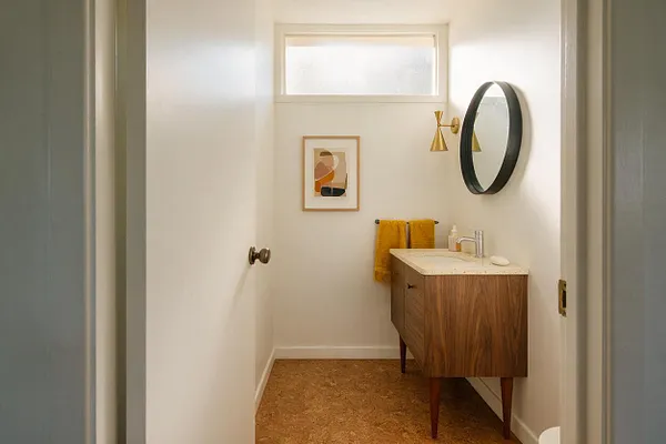 A compact powder room with white walls, a small horizontal frosted window, and a walnut vanity topped with a light stone counter. The space includes a round black-framed mirror, brass wall sconces, mustard towels, and a cork-colored floor for a warm mid-century modern look.