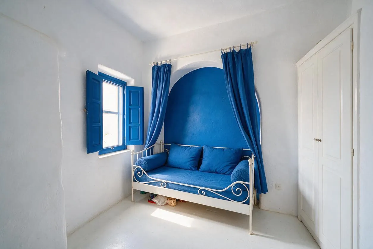 A bright kids' room with white plaster walls, a vivid blue alcove, matching blue curtains, and an iron daybed with blue cushions. A blue shuttered window brings in natural light, while a white double-door closet keeps the room simple and airy.