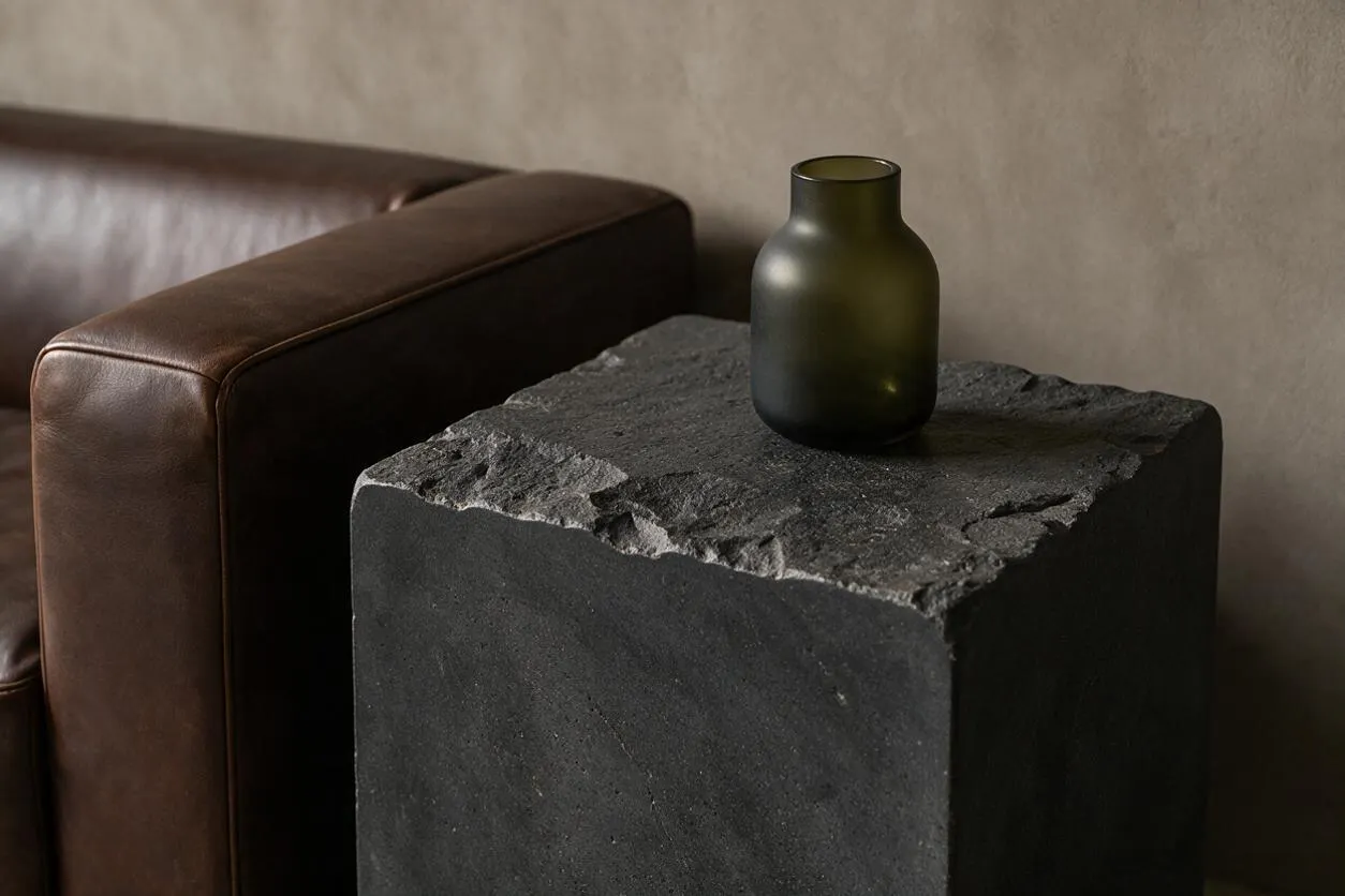 A brutalist-style interior vignette shows a rough, dark stone pedestal with an uneven chipped top surface and a small matte dark glass vase placed on it. A brown leather sofa arm appears to the left against a muted beige wall, creating a subdued, earthy composition.