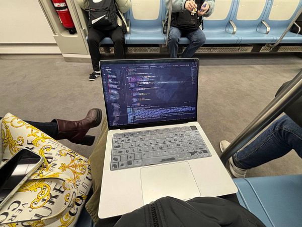 A person is working on a laptop while seated in a train, surrounded by other passengers.