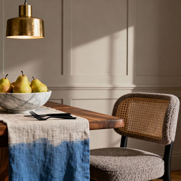Close-up of a transitional dining setting featuring a solid walnut table topped with a natural-and-blue dip-dyed linen runner. A white marble bowl filled with green pears and two matte black forks sit beneath a warm brass pendant. Behind, soft greige paneled walls catch daylight, casting gentle shadows. At the table is a cane-back chair with a bouclé-upholstered seat and rounded frame in black, blending traditional texture with contemporary lines.