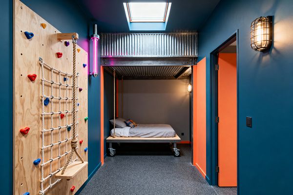 A kids’ room in industrial style features deep teal walls with bright orange doors, a central skylight, and caged wall sconces. A gray alcove holds a rope-suspended platform bed on casters beneath a corrugated metal canopy, while a plywood climbing wall with multicolored grips, a rope net, and a small rope swing lines the opposite wall; a neon pink tube light and speckled rubber flooring complete the playful, durable space.