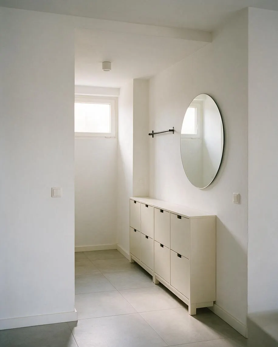 A bright minimalist entryway with white walls, a narrow cream-colored storage cabinet along the right wall, and a large oval wall mirror above it. The floor is light gray tile, and a small high window brings soft natural light into the simple, uncluttered space.