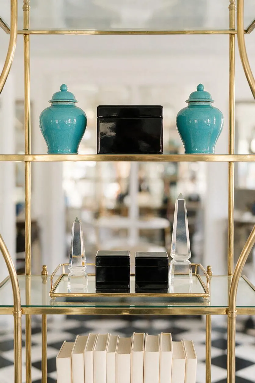 A Hollywood Regency-style etagere with a polished brass frame and glass shelves displays glossy turquoise jars, black lacquer boxes, clear crystal obelisks, and cream books. The blurred background and black-and-white checkered floor add depth and classic glamour.