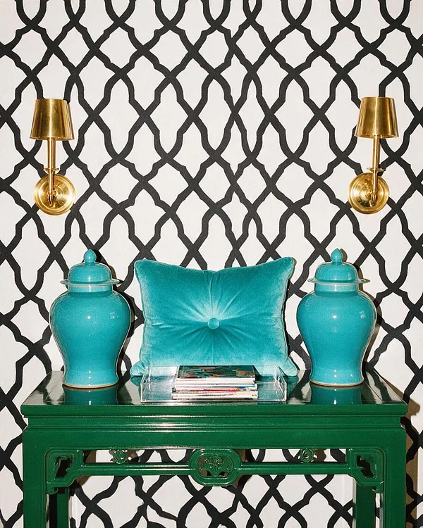 A Hollywood Regency-style vignette with a glossy emerald green console table against a white wall covered in bold black lattice-pattern wallpaper. Two brass wall sconces flank a turquoise velvet pillow, with matching turquoise ceramic ginger jars and a clear acrylic tray of magazines arranged on top.