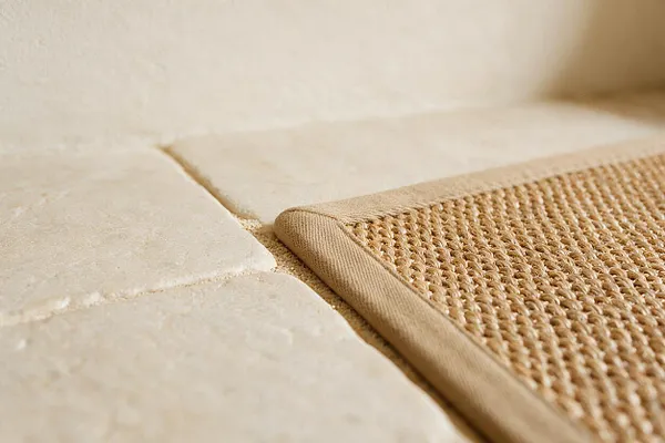 A close-up view of a beige woven mat with a bound edge resting on pale stone tiles. The scene highlights the contrast between the rough woven texture and the smooth, warm-toned stone surface, creating a calm Mediterranean-inspired look.