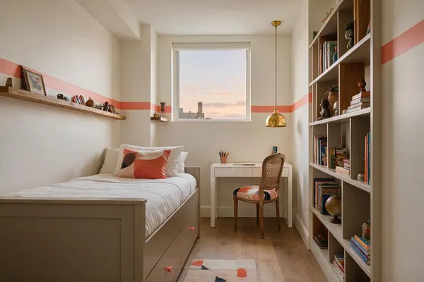 A narrow kids' room features a twin bed with white bedding, a coral-and-peach geometric pillow, a small white desk under a window, and a cane-back chair with a patterned seat cushion. Built-in shelving and a tall bookcase hold books and decorative objects, while a coral stripe wraps around the cream walls and warm wood flooring adds softness.