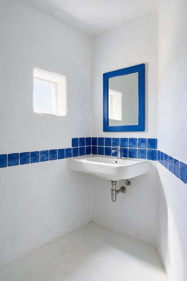 A small Mediterranean-style powder room with white stucco-like walls, a compact wall-mounted sink, and a bright cobalt blue mirror frame. A band of square blue tiles wraps around the room, and soft daylight enters through a small window, creating a clean, airy feel.