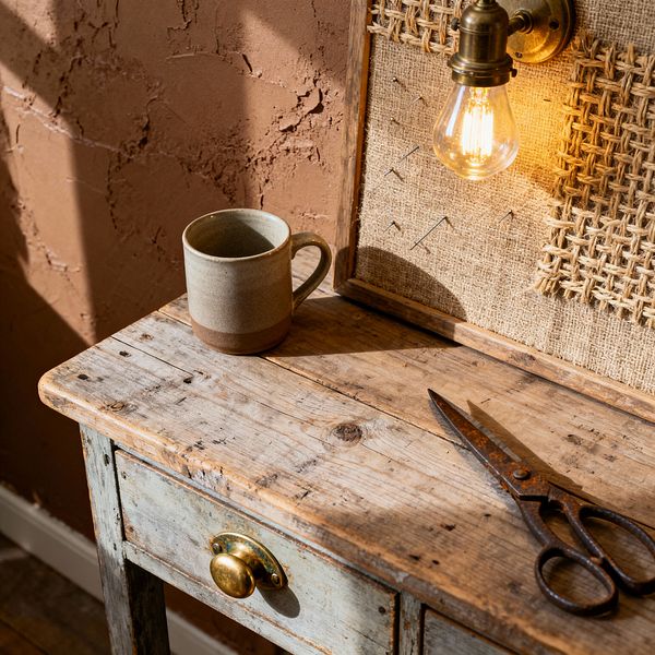 A close-up of a farmhouse home office vignette showing a weathered, reclaimed-wood desk with a brass cup pull. On the surface sit a two-tone stoneware mug and vintage iron scissors. A framed burlap pinboard rises behind the desk, fitted with an antique brass sconce and glowing exposed Edison bulb. The background is a rough terracotta plaster wall with sunlit shadows, creating a warm, rustic atmosphere.