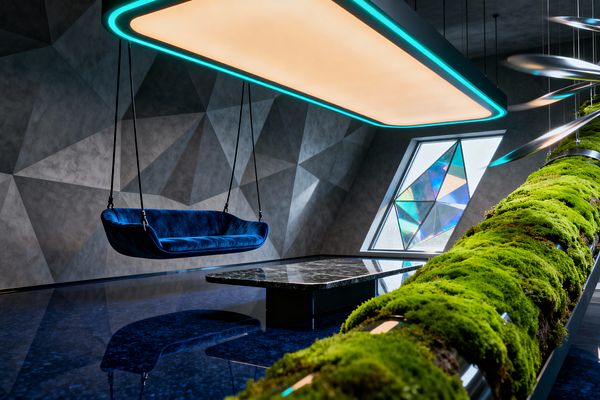 A futuristic living room features a cobalt-blue velvet swing sofa suspended by black cables beneath a large rectangular ceiling light with a warm glow and cyan edges. Faceted gray walls frame an angular window with iridescent glass. A low black marble coffee table sits on a glossy deep-blue floor that reflects the lighting. On the right, a chrome tube wrapped in bright green moss and thin hanging metal discs adds a biophilic accent.