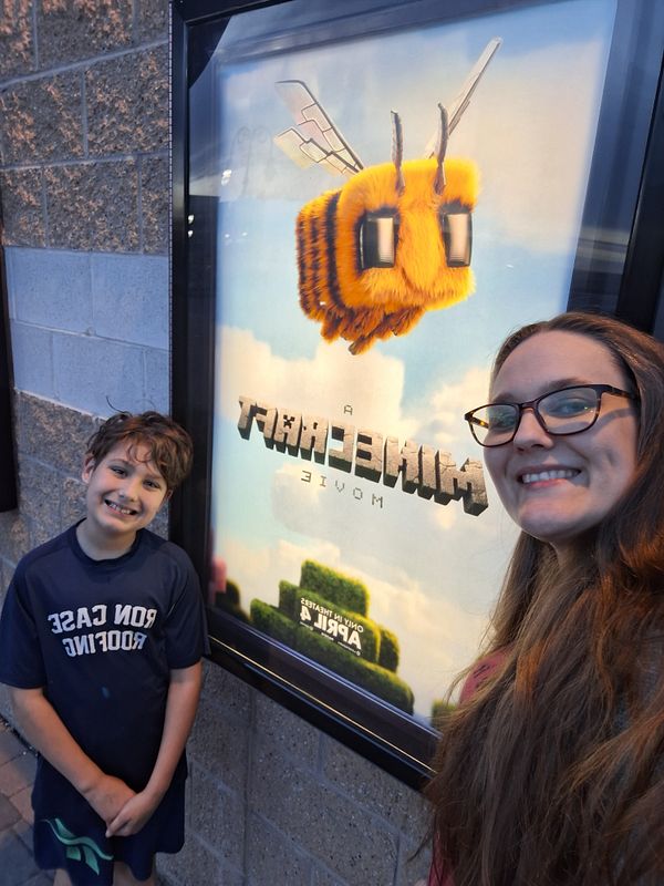 A woman and a boy pose in front of a Minecraft movie poster outside a cinema.