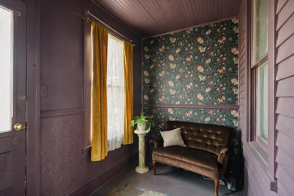 A small Victorian front porch with deep plum-painted wood walls, a dark stained ceiling, and floral wallpaper on the back wall. Mustard curtains frame a lace-covered window beside a tufted brown velvet settee, with a white pedestal holding a potted plant adding a decorative touch.