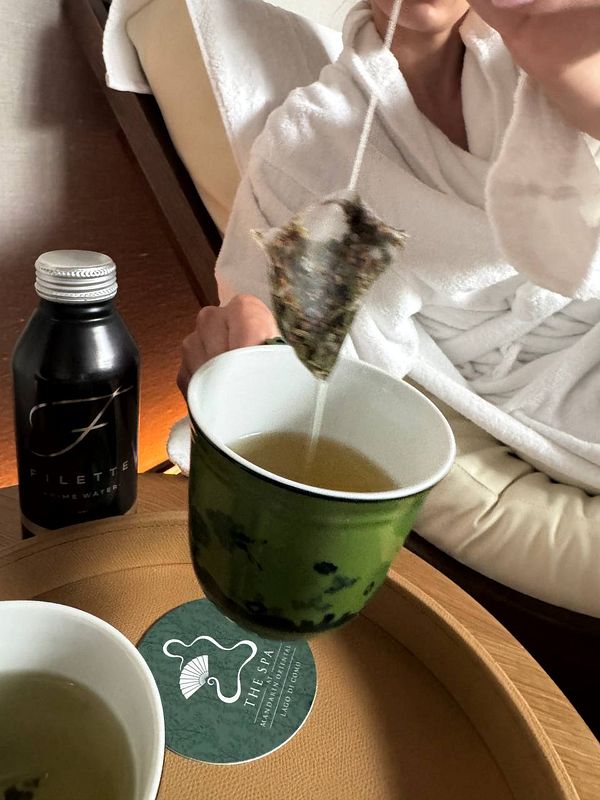 A person in a robe is enjoying tea from a microplastics-filled tea bag, with a bottle of water and a spa logo visible.