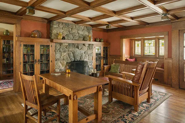 A Craftsman-style home office with a coffered wood ceiling, a large stone fireplace, built-in glass-front bookshelves, and warm wood trim throughout. The room includes a wooden desk, slatted wood chairs, a bench seating nook by stained-glass windows, and layered rugs over hardwood floors.