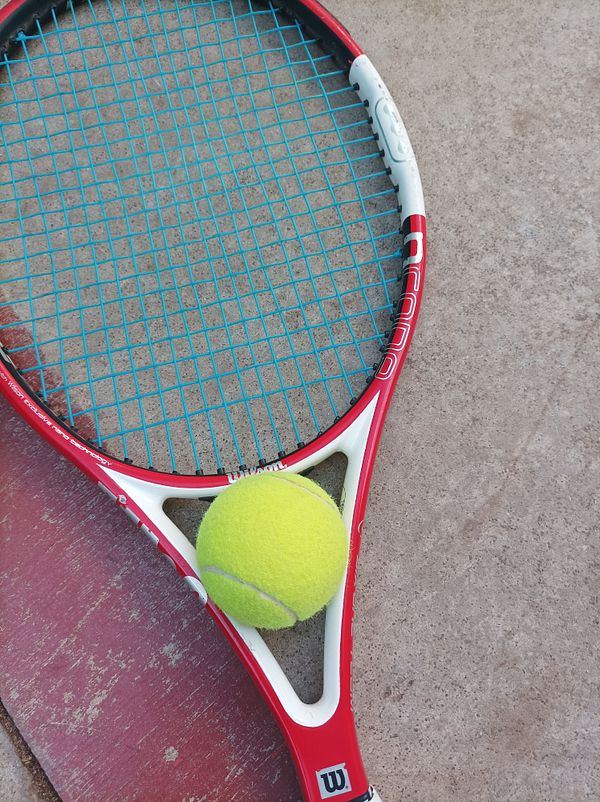 A tennis racket rests on a concrete surface with a bright yellow tennis ball positioned on its strings.