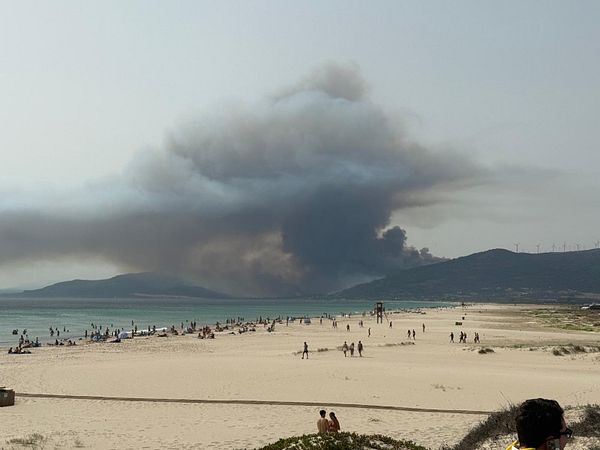 A large plume of smoke rises from a fire in the distance, visible from a beach crowded with people.