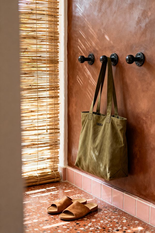 A compact mudroom corner shows a terracotta-toned plaster wall with three matte black iron pegs, one holding an olive canvas tote. A blush border of square tiles meets a warm red terrazzo floor flecked with white and charcoal. A rolled bamboo reed blind filters sunlight, casting linear shadows across the wall, bag, and floor, while a narrow strip of white plaster trim frames the edge; tan slide sandals rest neatly on the terrazzo.