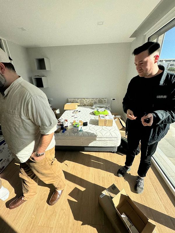 Two individuals are in a bright, sparsely furnished room with a bed and various items scattered around.