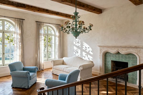 A sunlit French Country living room features arched windows with sheer linen curtains, exposed wood ceiling beams, and limewashed walls, with a cream linen sofa and two powder blue armchairs on an oak herringbone floor facing a carved limestone fireplace lined with green ceramic tiles. A patinated sage-green floral chandelier hangs above, casting leafy shadows on the wall, while a wooden stair railing appears in the foreground.