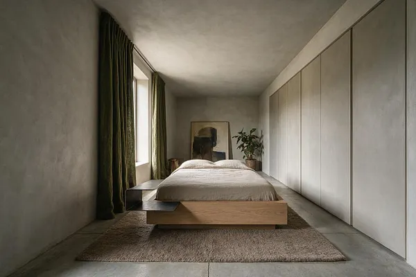 A long, narrow master bedroom with smooth concrete-style walls and ceiling, a low platform bed in light wood, and a neutral bedspread. Dark green floor-length curtains frame a side window, while large built-in wardrobe panels line the right wall and a textured rug anchors the bed.