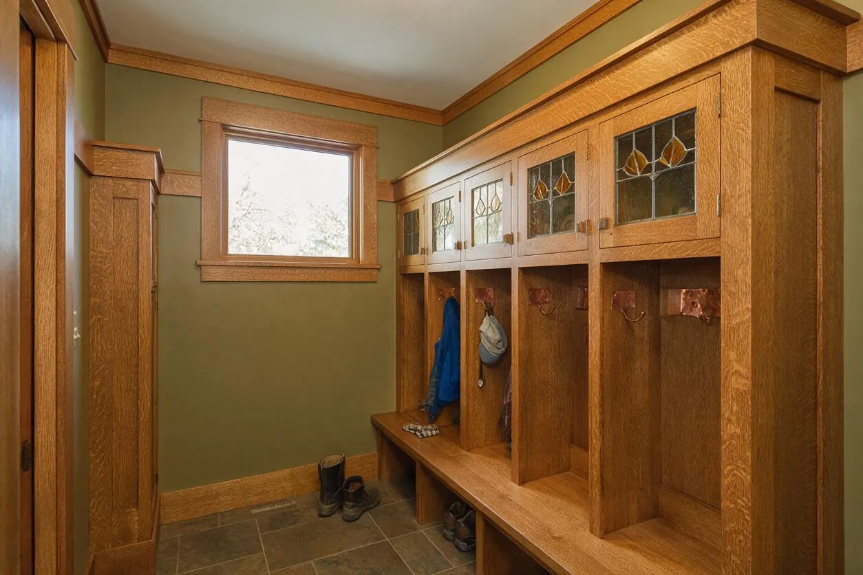 A Craftsman-style entryway with built-in oak storage cubbies and a bench lining one wall. The upper cabinet doors feature small leaded glass panels, while the lower open cubbies hold hooks for coats and bags; olive green walls and natural wood trim create a warm, traditional feel.