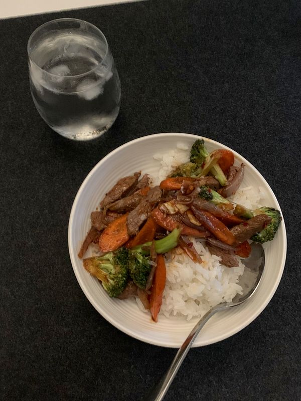 A bowl of deer stir-fry served over rice with a glass of water beside it.