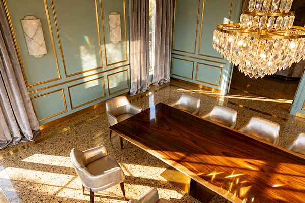 A sunlit dining room with pale sage paneled walls trimmed in gold, a large tiered crystal chandelier, and a polished walnut rectangular table on a brass plinth base. Champagne-upholstered armchairs with wooden legs surround the table, gray velvet floor-length curtains frame tall windows, terrazzo flooring with brass inlays gleams, and white marble wall sconces add sculptural accents for a vintage glam ambiance.