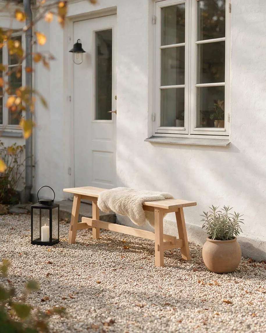 A serene Scandinavian-style exterior with a white stucco wall, cream-trimmed windows, and a pale wooden bench set on a gravel courtyard. A black lantern with a candle and a terracotta plant pot add simple, natural accents.