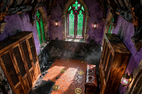 An atmospheric mudroom with Gothic details features pointed-arch stained glass windows in green and magenta set in stone, distressed purple walls, and dark wainscoting. The ceiling shows charred timber trusses, while the floor has hexagonal terracotta tiles dotted with brass medallions and a black grate; two tall wood lockers with lattice doors flank a brown tufted leather bench, and jewel-toned wall sconces add a moody glow with wisps of mist near the baseboards.