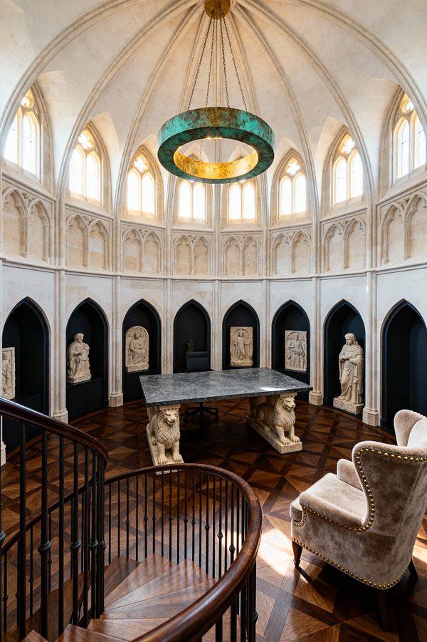 A circular Gothic home office features a ribbed vaulted ceiling and tall lancet windows that bathe creamy limestone walls in warm light. At the center, a charcoal stone desk on lion-carved pedestals stands on patterned walnut parquet; matte-black pointed-arch niches display statues and reliefs, while a taupe velvet wingback chair, a spiral stair with dark wood rail, and a suspended verdigris ring chandelier complete the dramatic space.