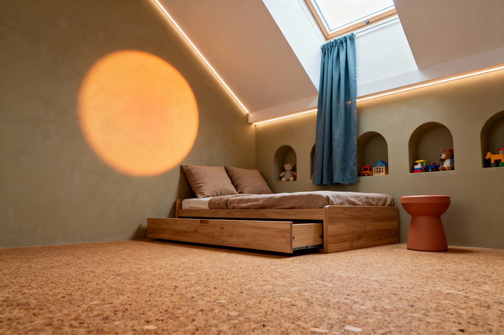 A minimalist children’s room with a low oak bed and pull-out trundle on cork flooring, set against sage plaster walls punctuated by arched niches displaying toys and a teddy bear. A roof skylight with a teal curtain and warm LED strip lighting illuminate the space, while a terracotta stool and a large projected orange circle on the wall add color to the neutral bedding and uncluttered layout.