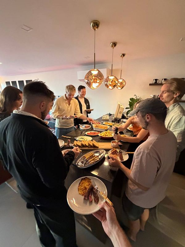 A group of people serving themselves food from a buffet-style spread in a modern kitchen setting during a coworking event.