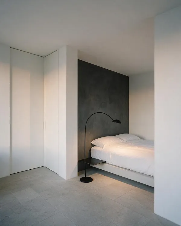A minimalist guest bedroom with a low white bed set against a dark charcoal accent wall, paired with a black arched floor lamp and a small floating bedside shelf. The room has smooth white walls, a clean built-in closet, and large light gray stone floor tiles that create a calm, spare atmosphere.