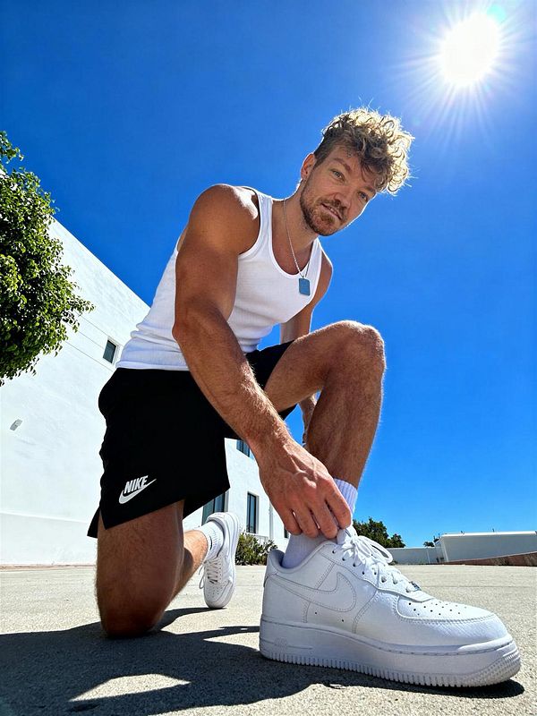 A man kneels on a sunny day, tying his white Nike sneakers while wearing a tank top and shorts.