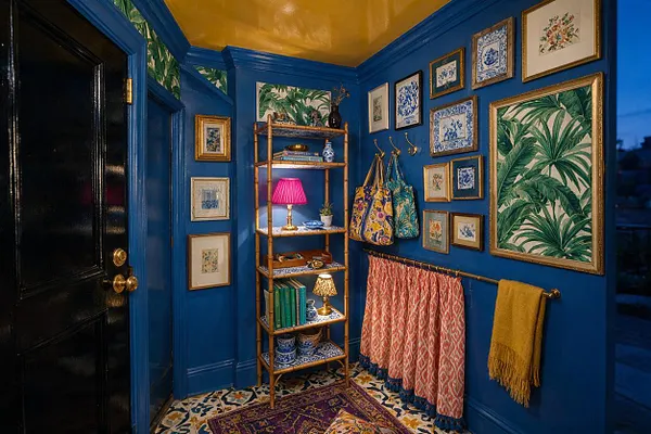 A richly decorated entryway features vivid cobalt blue walls, a glossy black door with brass hardware, and a gold-painted ceiling. The space is filled with framed botanical and floral art, a bamboo-style shelving unit, patterned textiles, hanging tote bags, and a colorful tiled floor with layered rugs, creating a bold maximalist look.