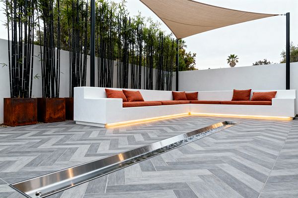 A modern patio with an L-shaped white built-in bench fitted with rust-colored cushions and underlit by warm LED strips sits on gray herringbone porcelain pavers. A narrow stainless steel linear water feature cuts through the floor, black bamboo in weathered corten steel planters lines a white privacy wall, and a beige triangular shade sail is stretched between black posts overhead, creating a minimalist outdoor lounge with a restrained palette.