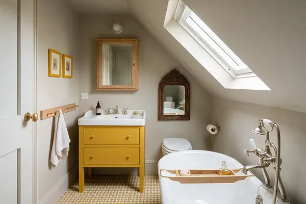 A compact attic bathroom with sloped ceilings, a large skylight, and a bright mustard yellow vanity topped with a white sink. The room includes a white freestanding tub with vintage-style silver fixtures, an ornate wall mirror, and patterned tile flooring in warm neutral tones.
