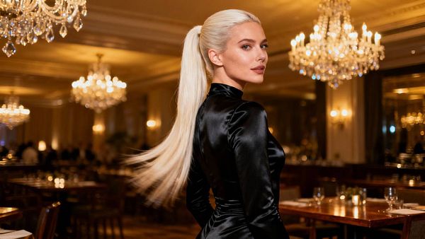 A stylish woman with long, straight blonde hair poses elegantly in a luxurious restaurant setting.
