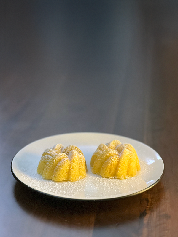 Two small, yellow cakes are presented on a white plate, dusted with powdered sugar.