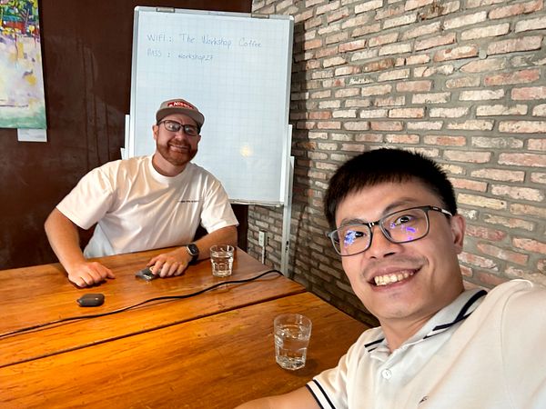 Two men are smiling and taking a selfie in a cozy café setting.