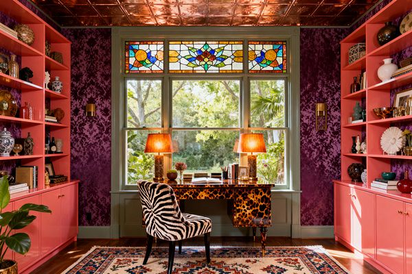 A maximalist home office with coral-pink built-in shelves packed with ceramics frames a large window topped by a colorful stained-glass transom. Deep purple flocked wallpaper, pressed copper ceiling tiles, a glossy tortoiseshell desk with two amber-shaded glass lamps, a zebra-print chair, brass sconces, hardwood floors, and a patterned rug create a layered, high-contrast workspace overlooking a lush garden.