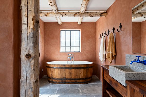 Rustic bathroom with terracotta plaster walls and a freestanding soaking tub clad in wood barrel staves, centered under a black‑grid window. The room features gray limestone floor tiles, birch log ceiling beams and a rough post, black wrought‑iron hooks holding neutral towels, and a wood vanity with a stone vessel sink and vivid blue wall‑mounted faucet, all lit by natural daylight.
